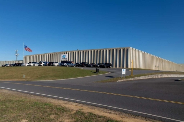 A large, beige industrial building with vertical siding sits behind parked cars and a U.S. flag under a clear blue sky. A sign and driveway are visible in the foreground, suggesting Kobayashi's expertise in product development workflow software.
