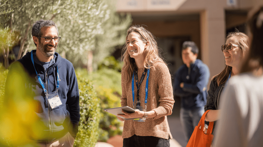 A group of people with conference badges smile and chat outdoors in a sunny setting, surrounded by greenery. One woman holds a tablet, and the atmosphere appears relaxed and friendly.
